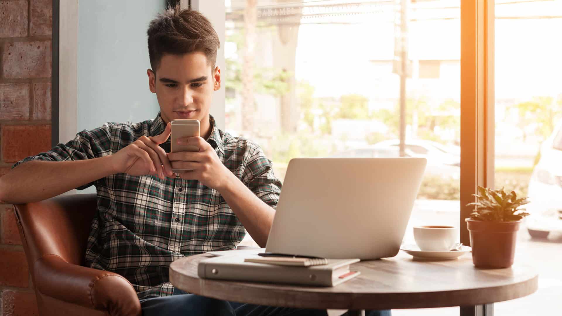 Jeune homme consultant son téléphone mobile avec un laptop, illustrant l'accès mobile aux services financiers