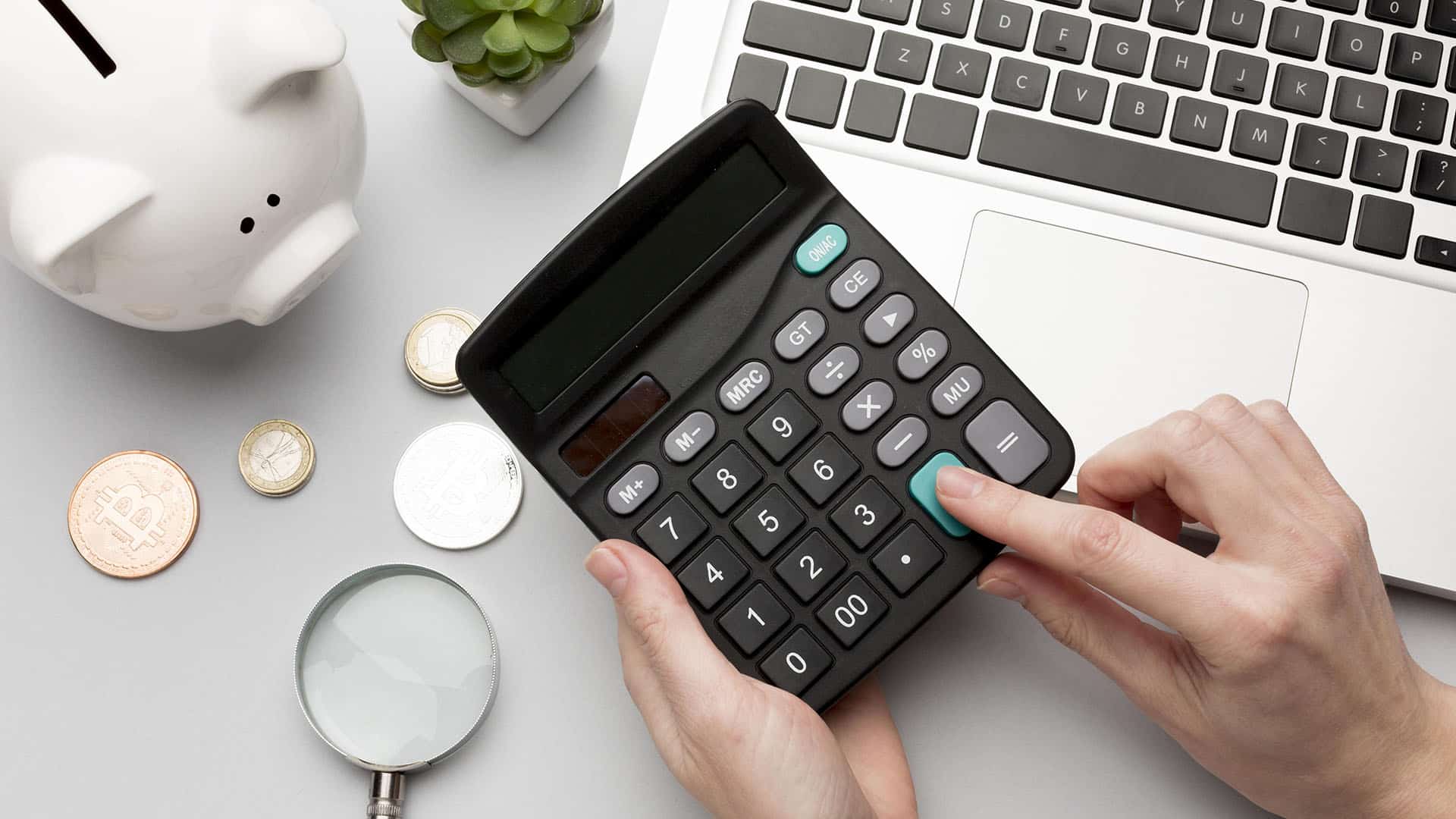 Main utilisant une calculatrice noire avec monnaies CHF, tirelire et clavier d'ordinateur sur un bureau blanc