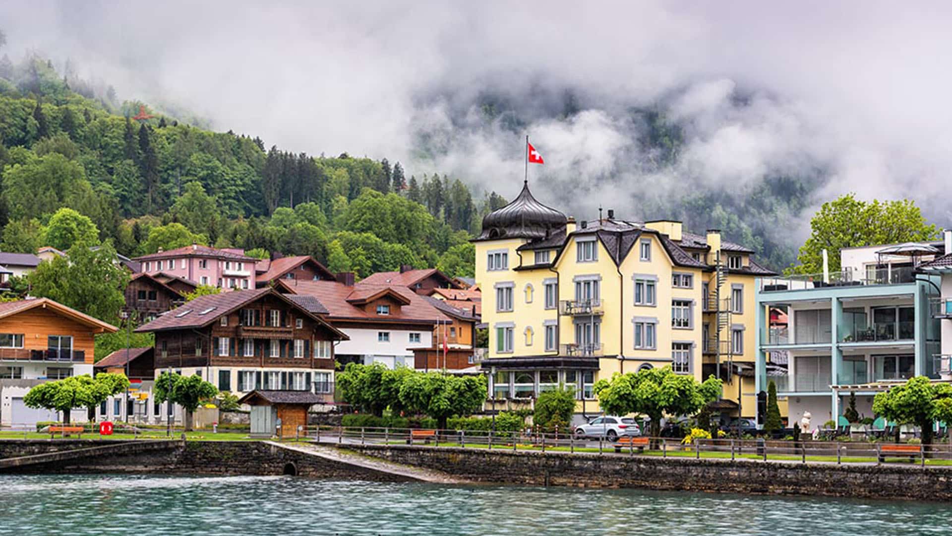 Village suisse au bord d'un lac avec bâtiments colorés, drapeau suisse et montagnes brouillées en arrière-plan