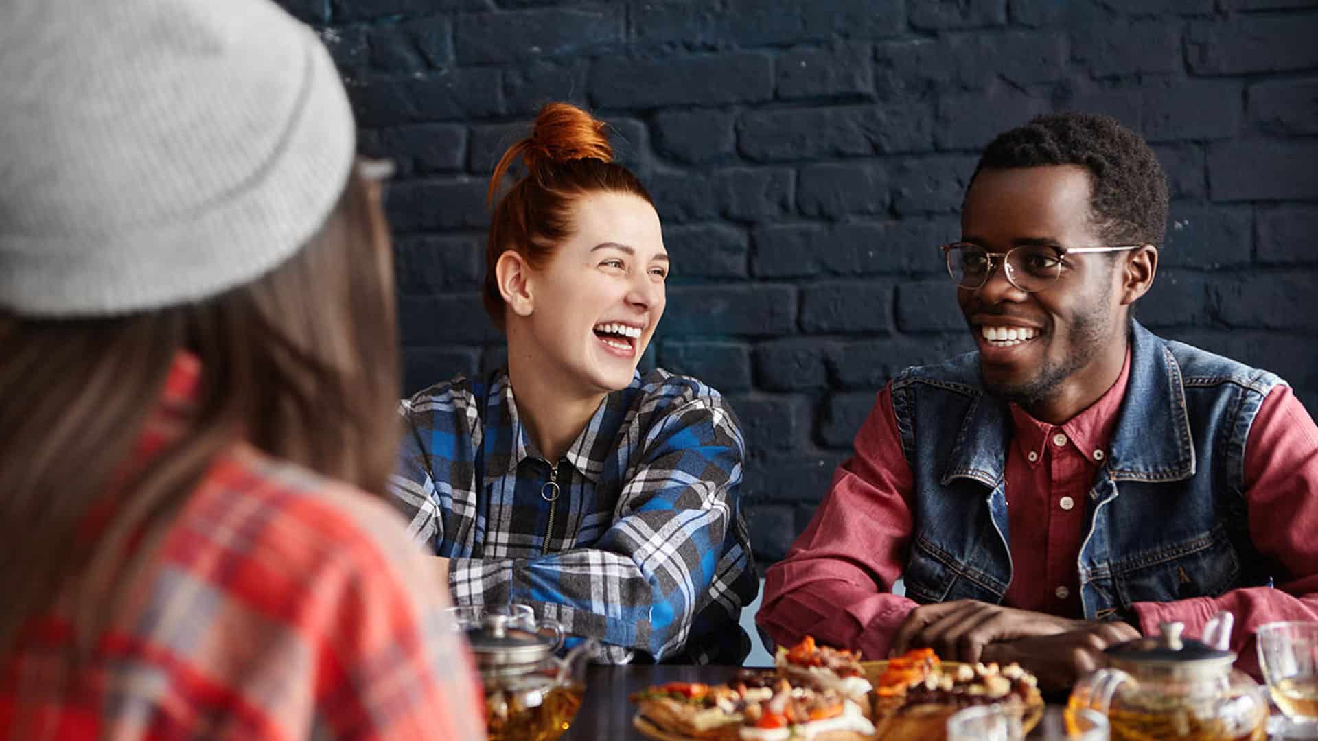 Trois amis souriants autour d'une table avec de la nourriture et des boissons, devant un mur en briques noires