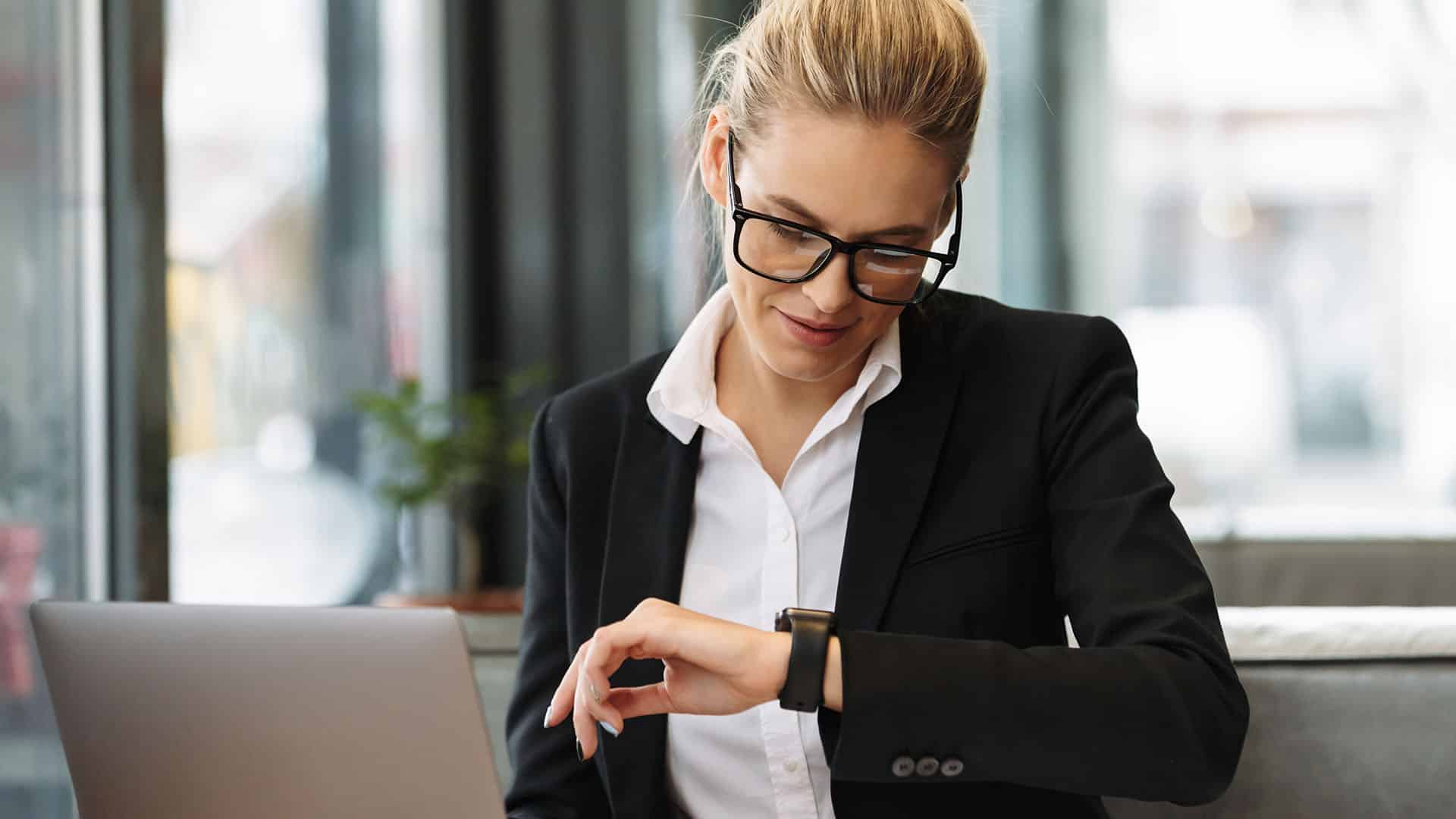 Femme d'affaires en costume noir vérifiant l'heure sur sa montre devant son ordinateur portable en bureau moderne