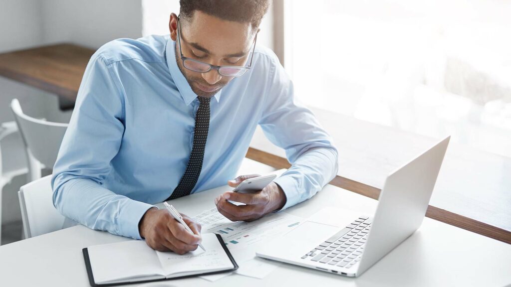 Homme d'affaires en chemise bleue analysant des documents financiers avec calculatrice et ordinateur portable