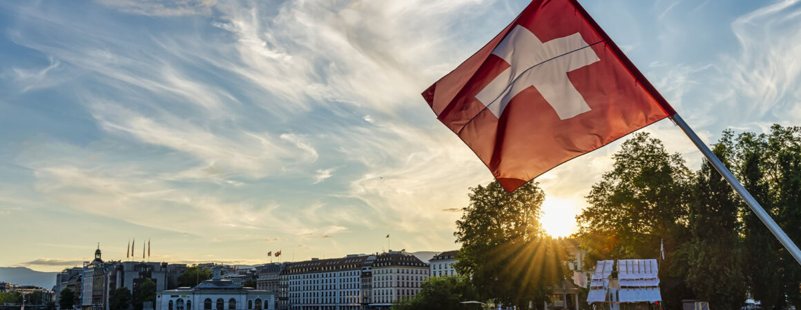 Drapeau suisse rouge et blanc flottant sur le Lac Léman avec la skyline de Genève en arrière-plan et coucher de soleil