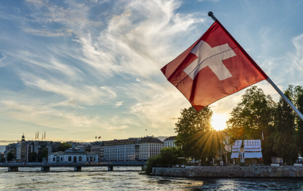Drapeau suisse rouge et blanc flottant sur le Lac Léman avec la skyline de Genève en arrière-plan et coucher de soleil