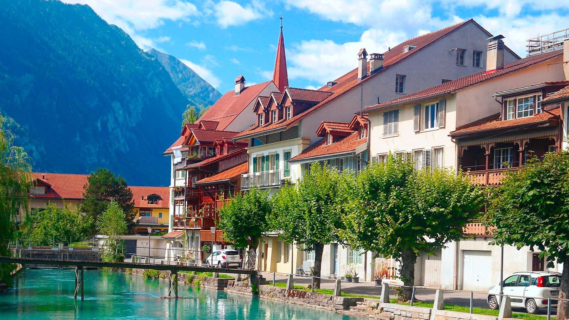 Village suisse avec maisons colorées, église au clocher rouge et rivière turquoise, montagnes en arrière-plan