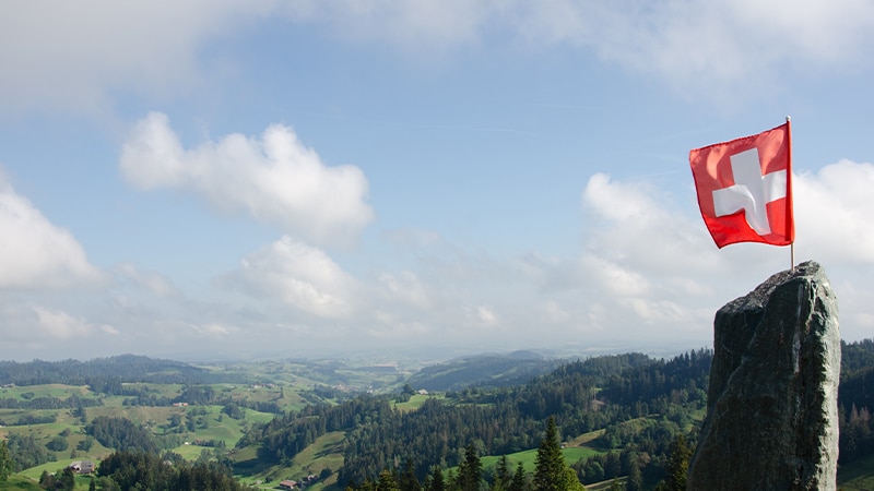 Drapeau suisse rouge et blanc flottant sur un rocher face aux montagnes et vallées vertes de Suisse
